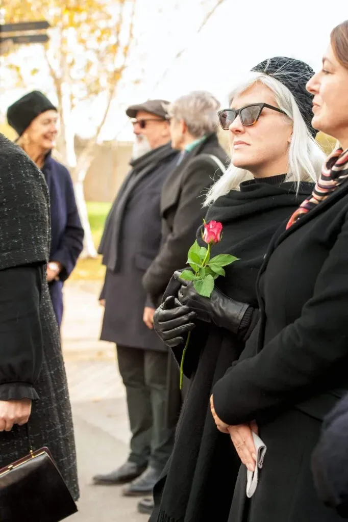 Femme vêtue de noir tenant une rose rose à la main lors d'un hommage funéraire en extérieur.